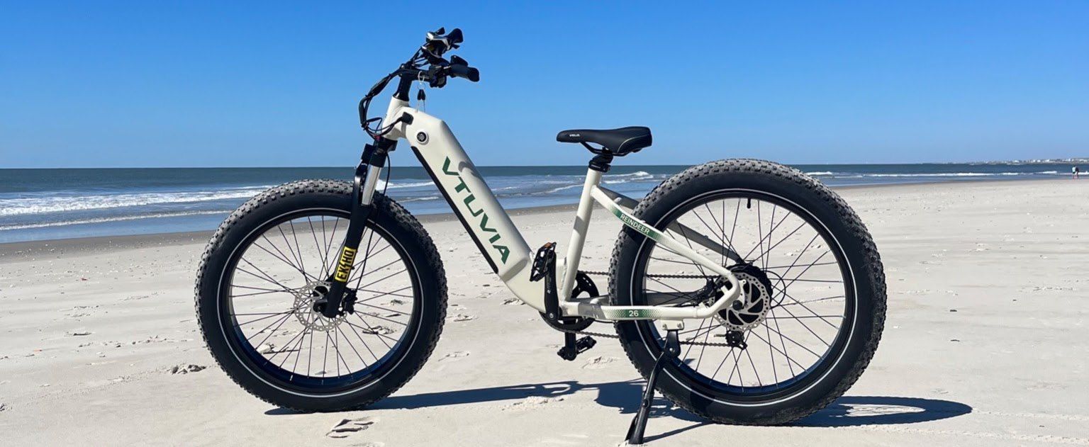Cream fat-tire step-through e-bike in side profile with the surf behind it